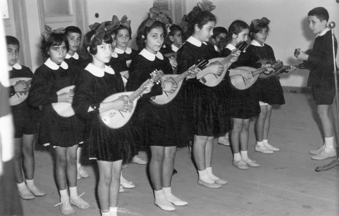 İlkokulumuzun “parlak” çocuklardan oluşan mandolin grubu (şef konumundaki ben): Erkek saçları alabros, önlükler koyu siyah, yakalar kolalı. Bu fotoğraf çekilmeden önce birkaç öğretmen bizleri, parmakların aynı perdeye basmasından ayaklarımızın aynı pozisyonda durmasına kadar, aynılaştırmak için epeyce uğraşmıştı. Duruş ve bakışlarımızdaki ürkekliğin nedeni odur. Çaldığımız parçalardan biri de “Neşeli ol ki genç kalasın” idi.